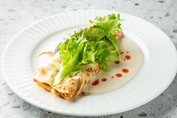 Rolled crepes filled with vegetables, topped with leafy greens and creamy sauce, served on a white plate with chili drops—gourmet presentation, vegetarian dish.