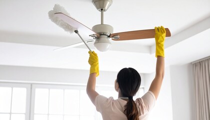 A woman in yellow gloves carefully cleans a ceiling fan with a duster in a bright room.