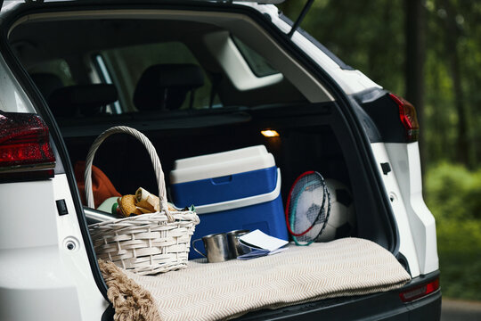 Open trunk of car showing picnic basket, cooler, badminton rackets, soccer ball, and travel gear packed for family road trip, outdoor forest background visible through trees