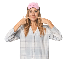 Blonde in pajamas with sleep mask smiles, pointing fingers at mouth.
