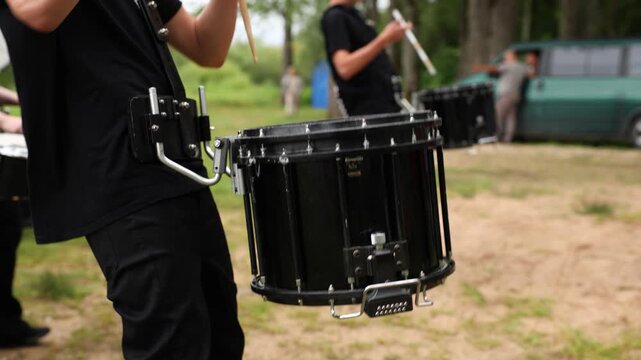 Close-up of a young drummer in black clothing playing a snare drum with drumsticks during an outdoor practice session with a marching band in a natural setting