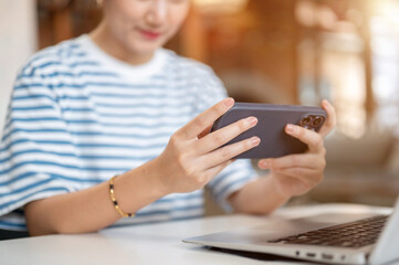 Close up of asian woman holding smartphone horizontal over laptop while sits at white table in cafe.