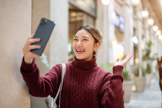 Young asian woman in knit sweater standing holding phone video calling in a shopping mall or market