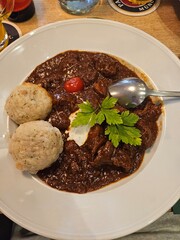 A close-up, top-down view of a plate of goulash with two dumplings, a dollop of sour cream, and a sprig of parsley, served with a spoon.