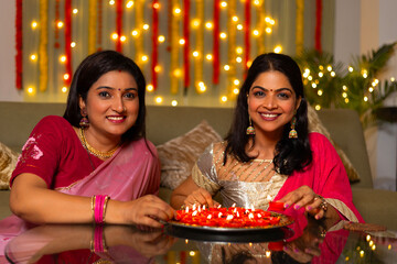 Indian beautiful women decorating together a thali full of diyas with smile on the occasion of Diwali season in colorful decorated home background - Diwali holidays festive