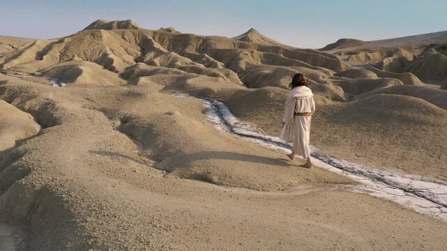 A mysterious man with long hair dressed in white, climbing the sandy hills of the desert. Side tracking shot
