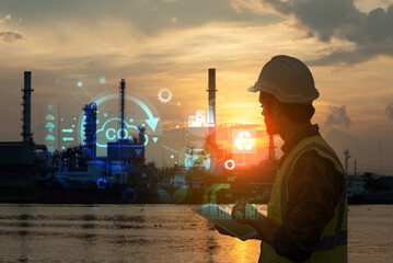 Engineer with safety gear using a tablet to monitor refinery operations enhanced with digital data graphics at sunset. Concept of smart industry and environmental technology.