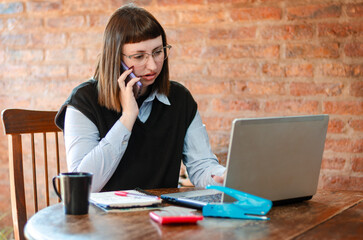 Young female employee working from home office with a laptop and talking on the phone looks...