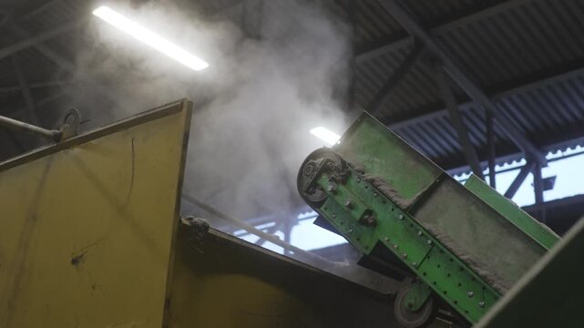 Shredded rubber drops steadily from tall conveyor into metallic container. Loose pieces scatter noisily below awaiting further steps