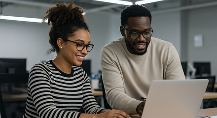 Two smiling Black professionals collaborating on a project using a laptop in a modern, open-plan office.