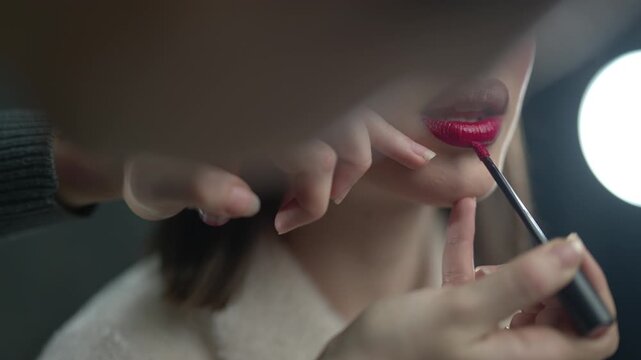 A makeup artist applies red lipstick to a model's lips in a professional setting. The model has dramatic eye makeup and a stylish haircut.