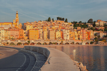View of the vibrant city with colorful buildings cascading down to the sea, a serpentine road hugging the coast, bathed in the warm glow of the morning sun, Menton, Alpes-Maritimes, France.
