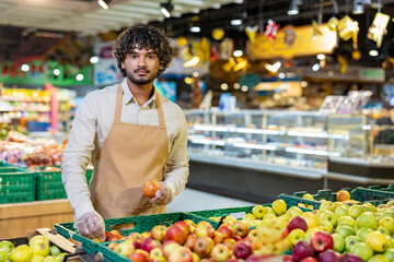 A supermarket worker in an apron holding an apple in front of a fruit display.