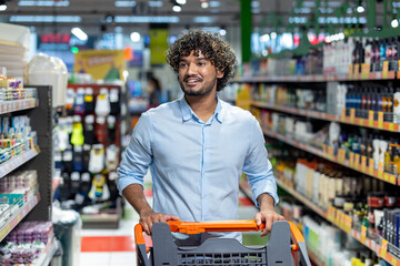 A man with curly hair smiles while pushing a shopping cart through a brightly lit grocery store aisle.