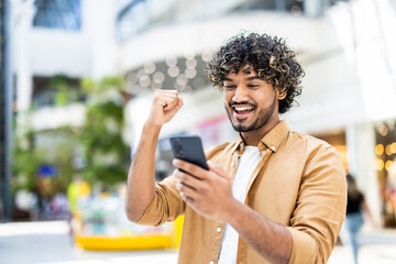 An excited young man with curly hair celebrates good news on his smartphone, smiling with delight in an urban setting.