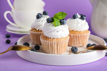 Plate of tasty cupcakes with blueberries on lilac background