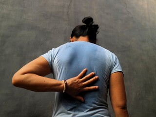 Asian Indonesian woman with stain on t-shirt sweat patch under back against gray background. Need deodorant. Uncomfort after exercise or hard activity
