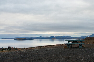 A fiery summer sunset with cloudy skies casts a vibrant glow over the calm waters of the lake and the distant shoreline with an empty bench