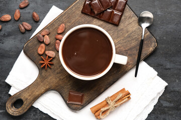 Wooden board with cup of tasty hot chocolate, cocoa beans and cinnamon on black grunge background
