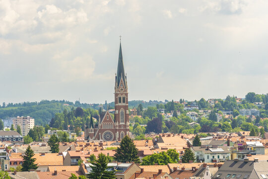 View of Herz Jesu Kirche soaring above a tapestry of red-tiled roofs, nestled amidst lush greenery under a soft sky, Herz jesu kirche church, Graz, Austria.