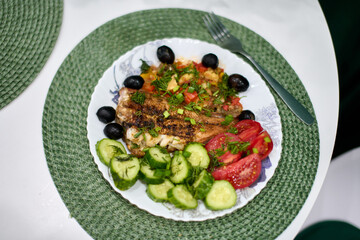 Grilled fish with vibrant vegetables and olives served on white plate. Decorative green mat adds texture. Bright natural light enhances fresh ingredients colors