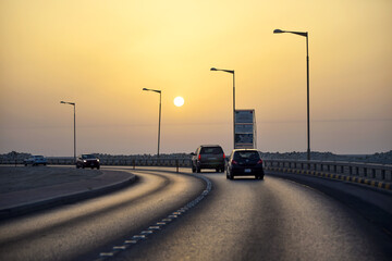 Manama, Bahrain - 29 May 2022: View of cars glide along a curved road at sunset, the sky a gradient of warm yellows and oranges above the Al Fateh Highway.