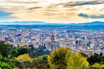 Go to Page
|12345...45Next
Panoramic aerial view of Girona with the Cathedral of Saint Mary rising above autumn foliage and urban rooftops. Distant blue mountains and dramatic clouds complete the scen