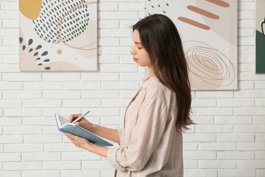 Young woman writing in notebook near paintings on white brick wall at art gallery exhibition