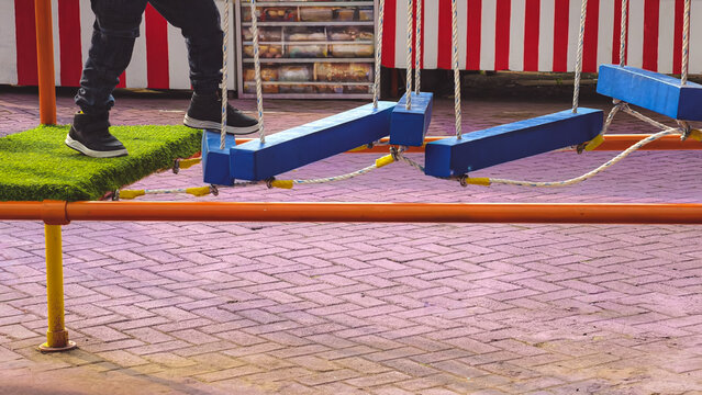 A child's legs walk across a colorful, outdoor playground structure with blue planks and ropes.