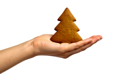 A close-up of an open palm holding a gingerbread cookie in the shape of a Christmas tree. The palm is positioned so that it appears to be offering or holding the cookie. The cookie is a golden brown c