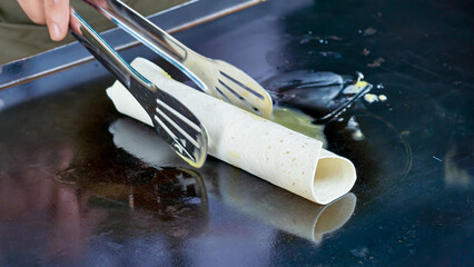 A hand uses tongs to cook a rolled crepe on a hot griddle.  The crepe is light-colored and appears...