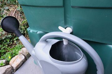 Grey watering can being filled from a rainwater tank tap, surrounded by green foliage and rocks, in...