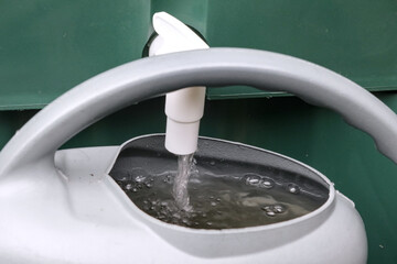 Grey watering can being filled from a rainwater tank tap, surrounded by green foliage and rocks, in...