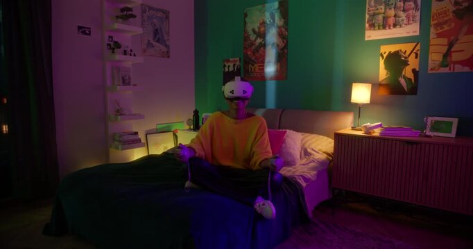 Young Black Woman Sitting on a Bed in a Colorful Neon-Lit Room at Home, Wearing a Virtual Reality Headset, Using Controllers with Her Hands while Exploring a 3D Interactive VR Experience