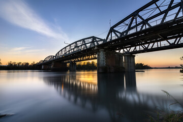 The Iron Bridge of Cremona spanning the Po Rover at sunset. Warm golden light casts long shadows on the structure, while the calm river reflects the vibrant colors of the sky