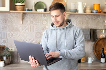 Young man chat with friends online from home holds a laptop in your hands. Smiling guy stand near kitchen desk read email message on laptop screen. Bearded freelancer male remote working from home.	