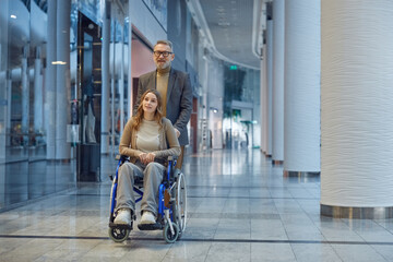 A man gently pushes a woman in a wheelchair down a long hallway © Nomad_Soul