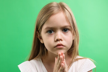 Little girl with clasped hands apologizing on green background, closeup