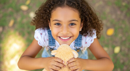 A smiling girl with curly hair holding a stuffed animal looking up at the camera outdoors in the park