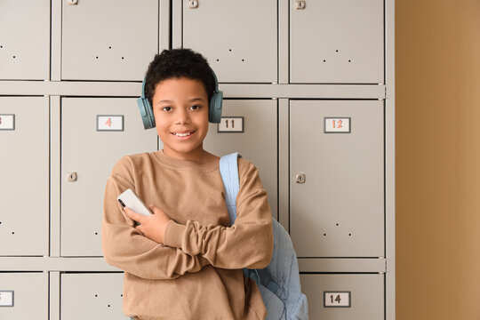 Teenage African-American schoolboy in headphones with mobile phone near locker on beige background