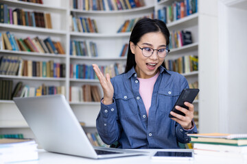 An Asian woman in a library is excited while looking at her phone. A laptop, tablet, and books sit on the desk.