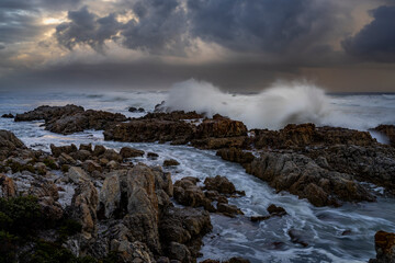 Powerful waves crash onto rugged rocks under dramatic storm clouds along the Hermanus coastline, Whale Coast, Overberg, Western Cape, South Africa — capturing the wild beauty of the seascape.