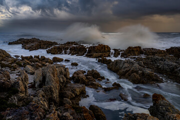 Powerful waves crash onto rugged rocks under dramatic storm clouds along the Hermanus coastline, Whale Coast, Overberg, Western Cape, South Africa — capturing the wild beauty of the seascape.
