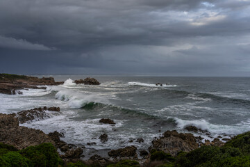 Powerful waves crash onto rugged rocks under dramatic storm clouds along the Hermanus coastline, Whale Coast, Overberg, Western Cape, South Africa — capturing the wild beauty of the seascape.