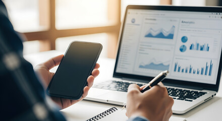 Person Analyzing Financial Data on Laptop and Smartphone in Natural Light Office Environment