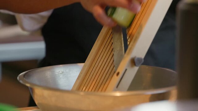 A chef slices a cucumber on a mandolin without using a safety guard, flipping it and turning it as he goes in an up and down motion.