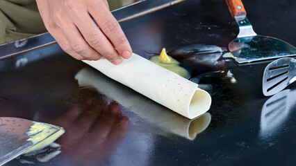 A hand prepares a tortilla wrap on a hot griddle, adding condiments.