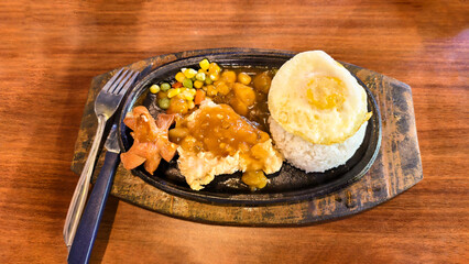 A wooden platter of a hot plate meal, featuring rice, fried egg, chicken, and vegetables.