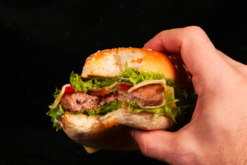 Hand holding a delicious burger with lettuce, cheese, and sesame bun on black background