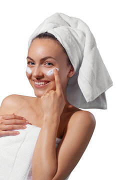 Smiling young woman applying facial cream under her eyes, wrapped in a white towel with a towel turban on her head